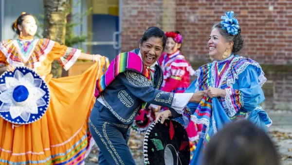 People are celebrating outside. They wear traditional Mexican dress and are smiling happily. In the foreground, one dancer is wearing a blue suit with a yellow decorative print following the seams, and a colourful striped garment over a shoulder. They are laughing and holding the hand of another dancer who is wearing a blue dress with a matching hair bow. The dress has lace trims and rainbow decoration at the collar and cuffs. They are holding a black wide-brimmed hat decorated with green and red. 