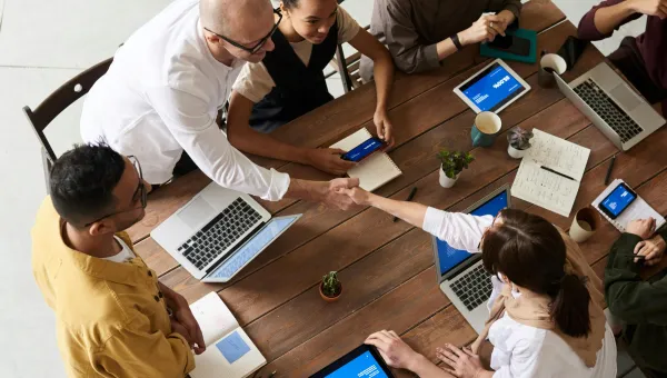 A top-down view of a diverse group of six professionals seated around a wooden table with laptops and tablets, engaged in discussion during a collaborative meeting in a modern workspace.