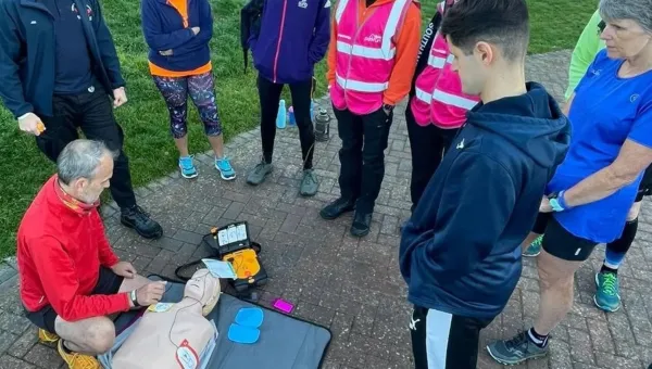 A group of people is observing a CPR training demonstration outdoors, with one person performing the demonstration on a training mannequin next to an AED device, in a sunny, grassy area.