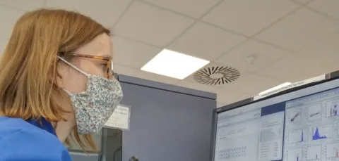 Anne Rogel in a blue lab coat and floral patterned face mask looking at a computer screen with scientific data in a lab