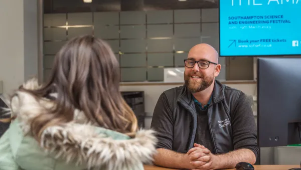 A University of Southampton staff member sits behind a desk while speaking with a student. The staff member is smiling and wearing University of Southampton-branded clothing.