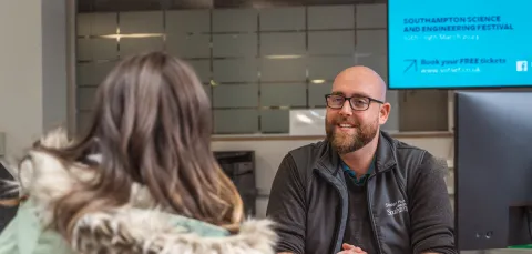 A University of Southampton staff member sits behind a desk while speaking with a student. The staff member is smiling and wearing University of Southampton-branded clothing.