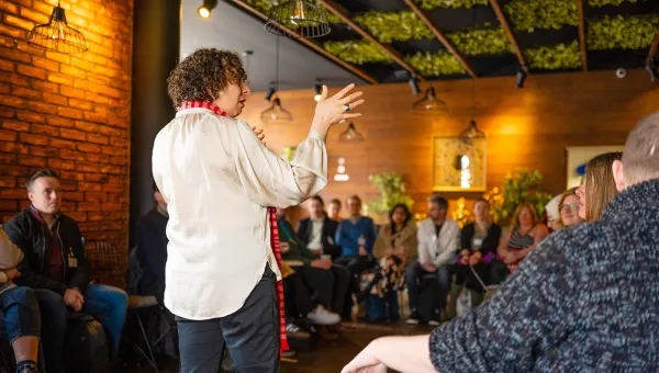 People are sitting around the edges of a room watching somebody giving a lively talk. 