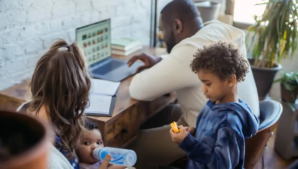 Family at home with father working on laptop, mother feeding baby, and young child playing nearby.