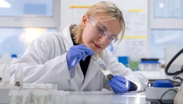 A researcher in a lab putting at a limpet shell in a test tube. The researcher is wearing a white coat, purple gloves and eye protection glasses.