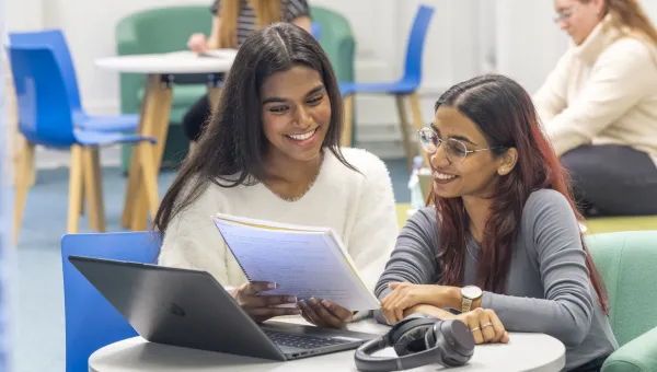 Two students huddle around a laptop together, whilst reading information on a large notepad.