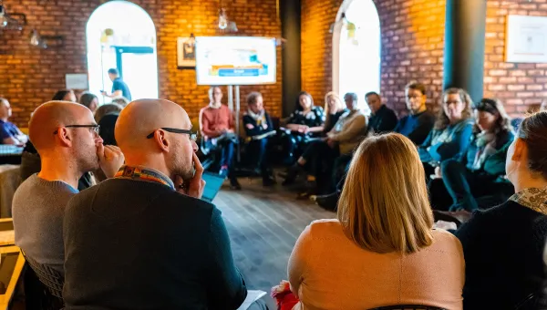 A group of people is seated in a circle. The participants are engaged in a discussion. A screen is visible in the background, displaying content