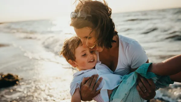 Woman holding a child on a sunny beach, both smiling.