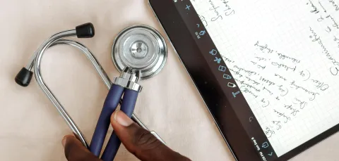 Hands write notes on a tablet with a stethoscope and a laptop near by.