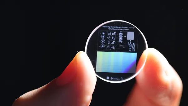 Zoomed in view of a hand holding a 5D memory crystal between two fingers. The memory crystal is a small clear disc with diagrams of human DNA on the surface.