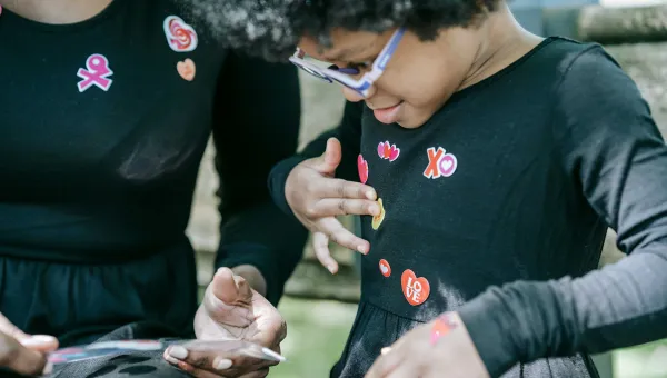 A young child is choosing stickers from a sheet held by an adult. The child is adding the colourful stickers to their top.  