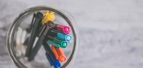 Top-down view of colourful pens in a glass jar on a grey wooden surface.