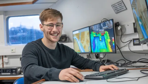 Daniel Hampton is sitting at the work station on Research Vessel Callista. There are screens displaying coulourful charts in the background.