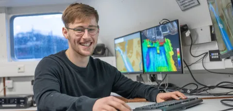 Daniel Hampton is sitting at the work station on Research Vessel Callista. There are screens displaying coulourful charts in the background.