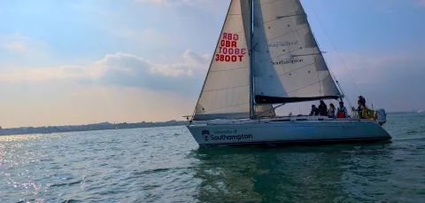 A yacht with 2 sails and a training crew sailing out on the water. The  sky has clouds and an orange eveningglowsky