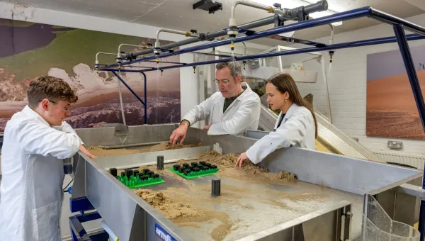 Three people in lab coats work at a sand-filled table with various objects and overhead nozzles, set against a backdrop of landscape images.