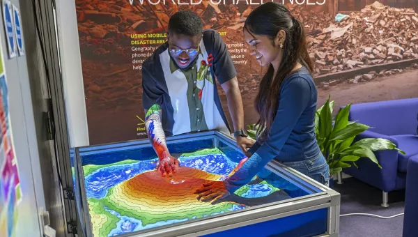 Two students exploring an interactive topographic sandbox display with projected contour and elevation colours.