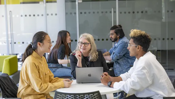 Three people sat a desk talking. The person in middle has a laptop open on the desk.