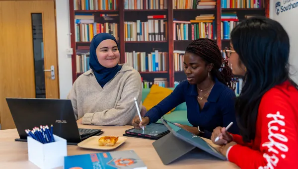 Nora and 2 friends study together in a library. They have their laptops open on the table in front of them.