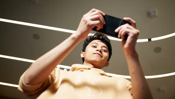 A student is looking down at the camera while using their smartphone, which they are holding horizontally. The student is wearing a pale yellow hoodie, and stood in a corridor which is well-lit by several fluorescent lighting strips.