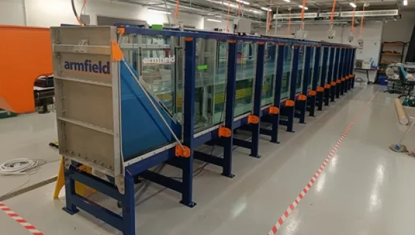 Long glass-sided wave tank in a laboratory, supported by a blue metal frame with orange fittings, and an Armfield wave generator positioned at one end.