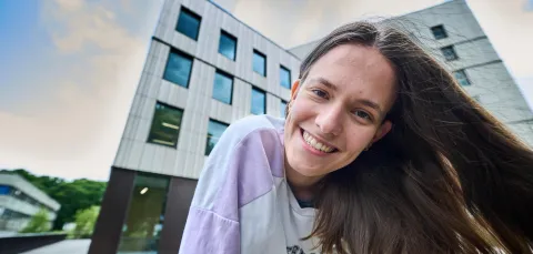 A smiling student stood outside the Centenary Building, also known as Building 100, on the University of Southampton's Highfield campus. They have long hair and wearing a purple and white shirt with blue jeans.