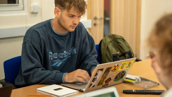 A student with short blonde hair sat at a desk with a laptop open. They are wearing a Reebok sweater, and several rings on their fingers.