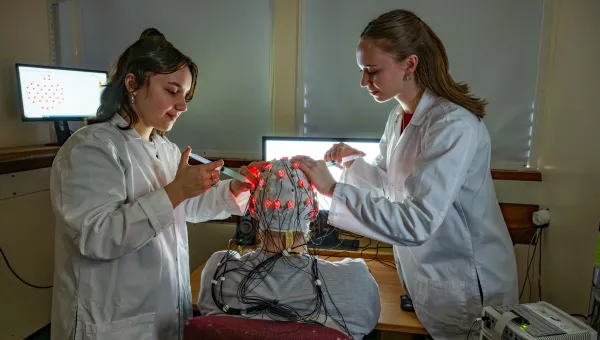 Two students adjust electrodes on a brain cap with red lights in a lab.
