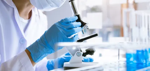A person in a lab coat using a microscope, wearing a face mask and blue gloves.