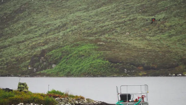 A drone hovers over a lake near a small floating platform with equipment, surrounded by lush green hills. The scene suggests a scientific or environmental study in a remote natural location.