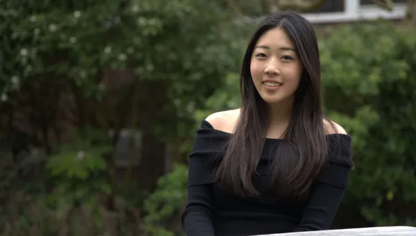 Student sitting outdoors with green shrubs behind her. She is smiling at the camera.