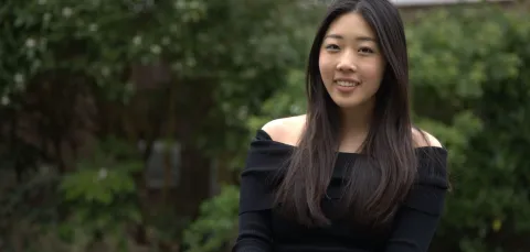 Student sitting outdoors with green shrubs behind her. She is smiling at the camera.
