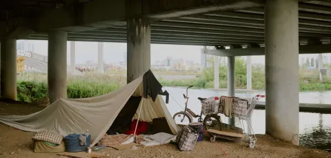 A makeshift shelter set up under a bridge near a river, with personal belongings, a bicycle, and a shopping trolley. The image reflects urban homelessness and temporary living conditions