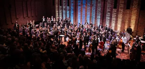 A full orchestra stands on stage in a concert hall with brick walls and vertical lighting accents, facing an audience seated in the foreground. Musicians hold various instruments including strings, woodwinds, brass, and percussion, with music stands arranged across the stage.