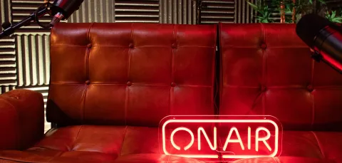 A red neon 'on air' sign on a brown leather sofa in a podcast recording studio, surrounded by two microphones set up on stands.