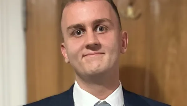 Daniel, a young man with brown hair and eyes and wearing a navy blue blazer and grey tie, smiles at the camera