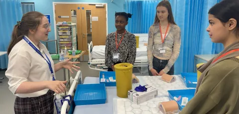 A group of students standing around a hospital bed, listening to a demonstrator.
