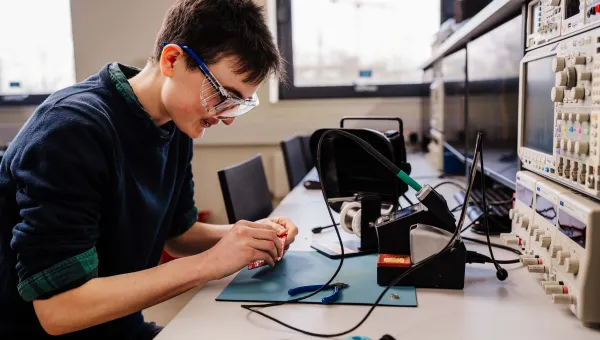 Biomedical engineering PhD student wearing safety goggles soldering a circuit board in an electronics lab.