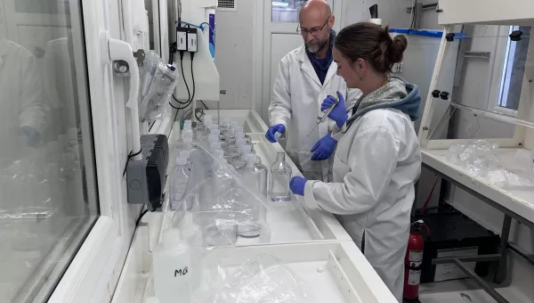 Two people in a laboratory preparing water samples, wearing lab coats and gloves, and handling bottles and equipment on a long workbench.