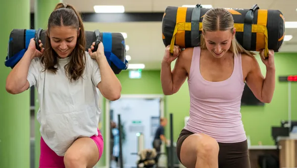 Two people in the gym, bending down on one knee. They have a weighted roll on their shoulders.