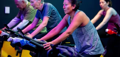 People taking part in an indoor cycling class, seated on stationary bikes in a dimly lit studio with colourful lighting.