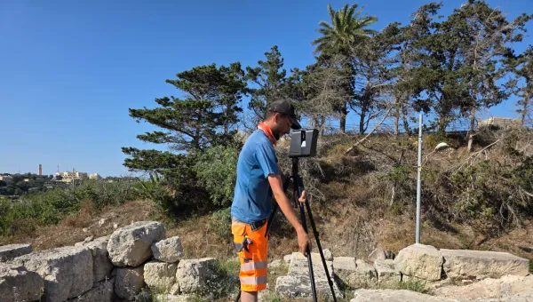 Ben, an archaeology and history BA student, uses surveying equipment on a rocky outcrop surrounded by trees in the mid-day sun