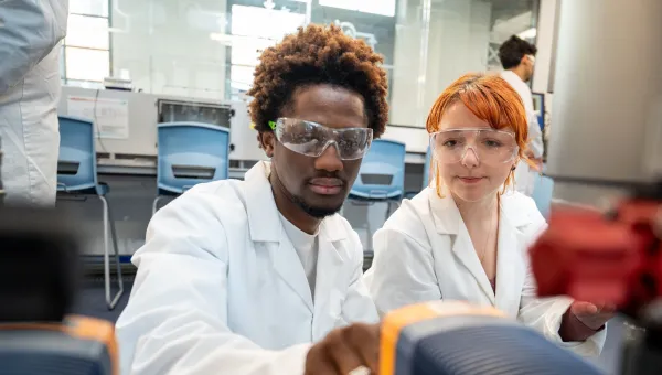 Two students wearing white lab coats and clear safety goggles are working together in a modern laboratory. The students are leaning over bench-top equipment adjusting a dial.