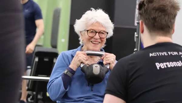 An elderly lady smiles at her instructor while lifting a kettle bell to her chest