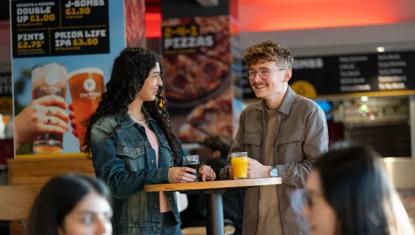 Two students chat over drinks in the Student Union. They're standing slightly to the side of each other, laughing together.