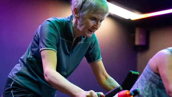 An elderly lady with short cropped hair rides a cycle machine during a class. The lights are low and moody and other participants focus on their session in the background