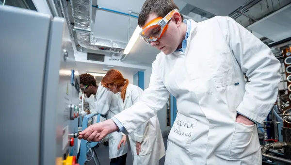Student in a chemical engineering lab wearing a white lab coat and safety goggles adjusts controls on industrial equipment, while two other students work at a panel in the background.