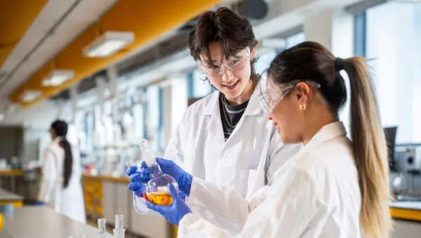 Two students in a laboratory wearing lab coats, safety glasses and gloves hold a glass flask containing orange liquid, with other lab equipment on a bench nearby.