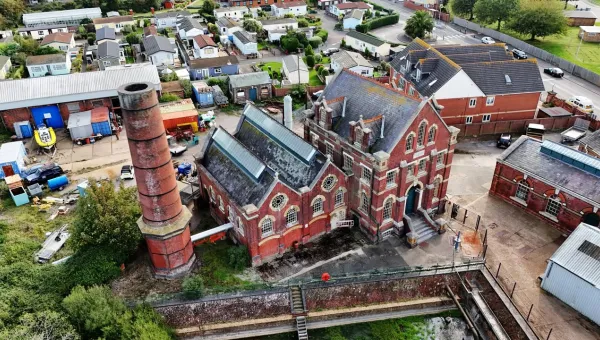 Aerial view of a historic red-brick industrial building with a tall chimney, surrounded by small homes, workshops, and a residential neighborhood.