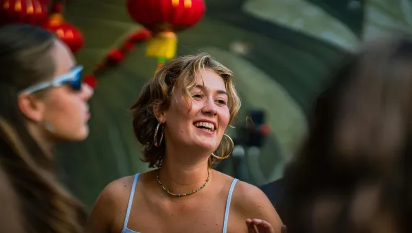 A close‑up shows a person wearing a white top and large hoop earrings, standing outdoors among others, with red lantern decorations hanging in the background. The scene appears to be a social gathering set in a narrow street with soft evening lighting.